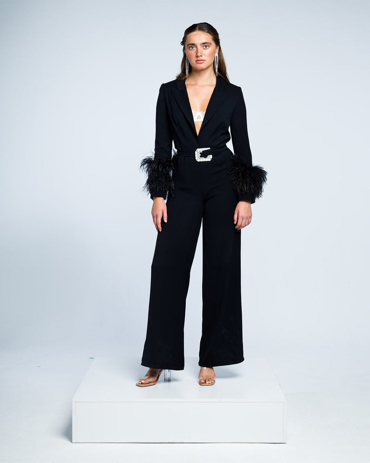 Woman in a black wide-leg jumpsuit with feather sleeve cuffs, pearl belt, and clear heels