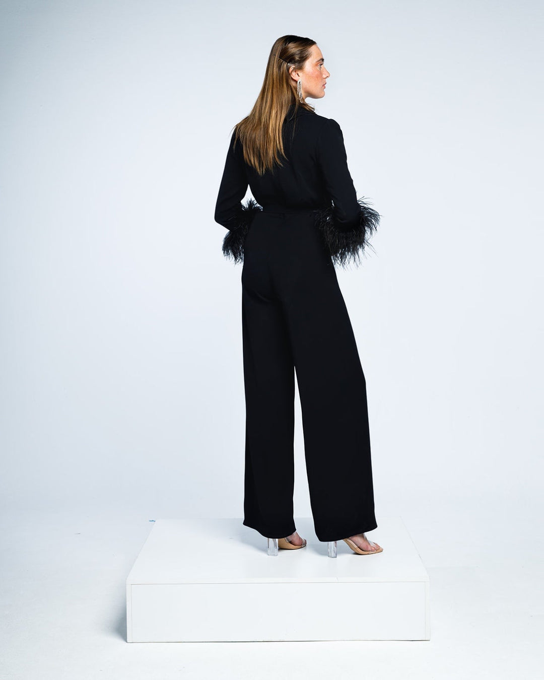 Woman modeling black jumpsuit with feathered cuffs and clear heels in studio