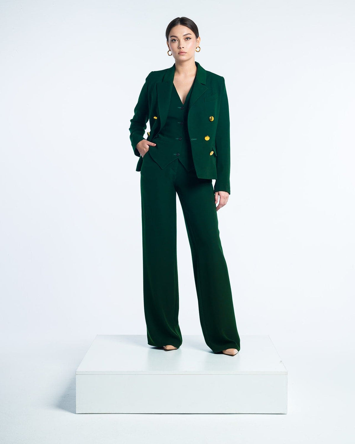 Woman in elegant dark green suit with gold buttons posing on a white platform studio
