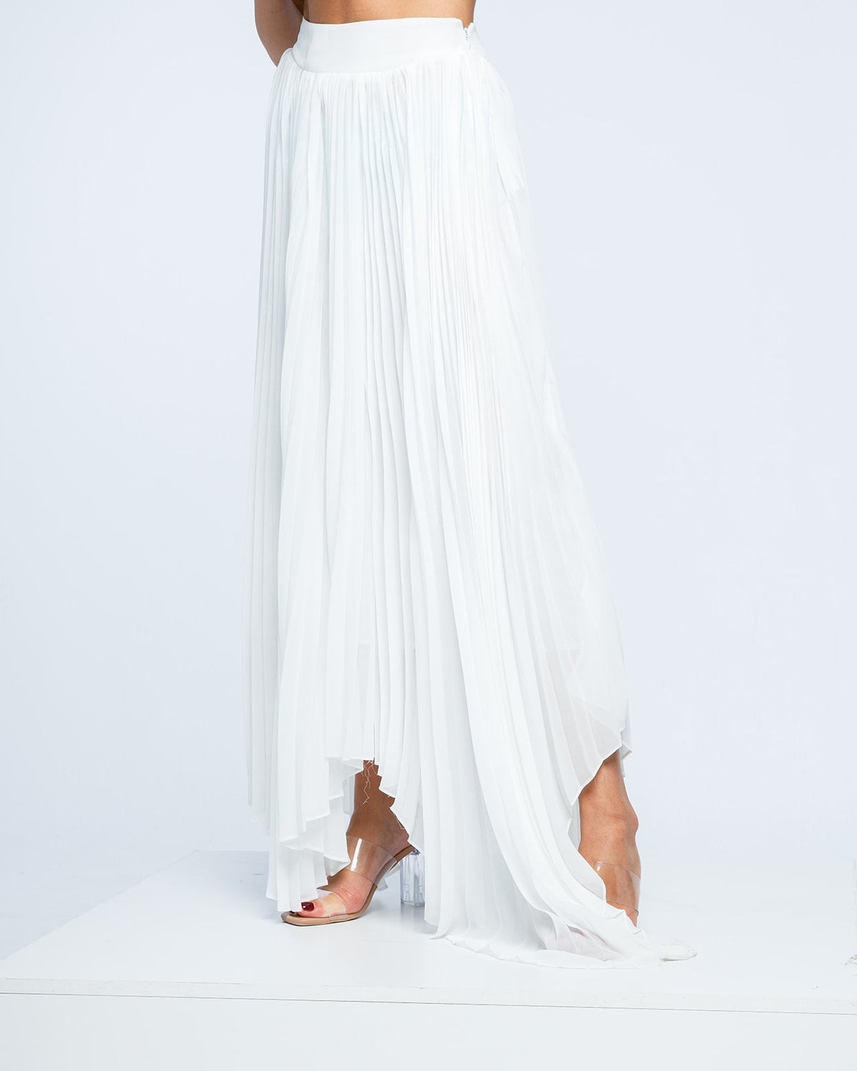 Woman wearing a long white pleated skirt and clear heeled sandals on white background