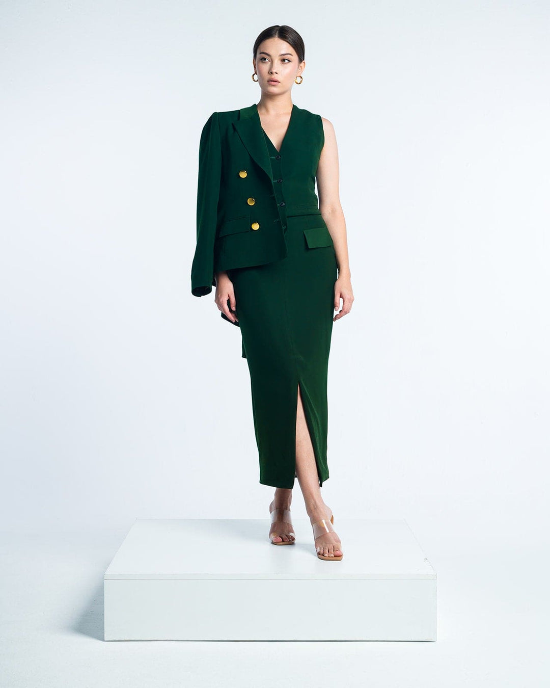Woman in elegant dark green sleeveless dress with gold buttons and asymmetric blazer, standing on white platform