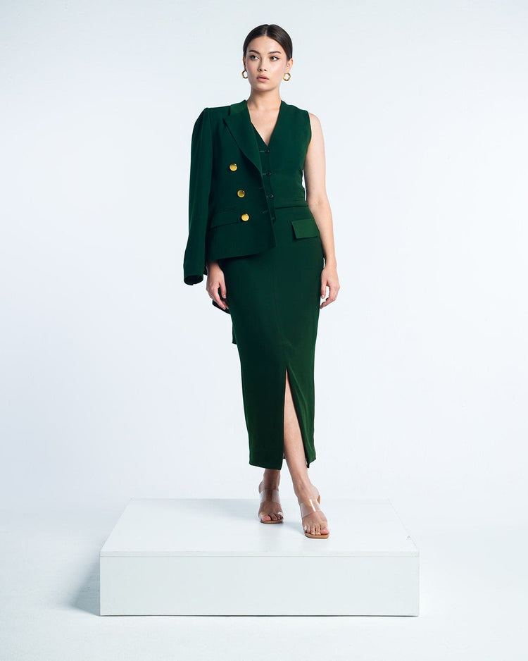 Woman in elegant dark green sleeveless dress with gold buttons and asymmetric blazer, standing on white platform
