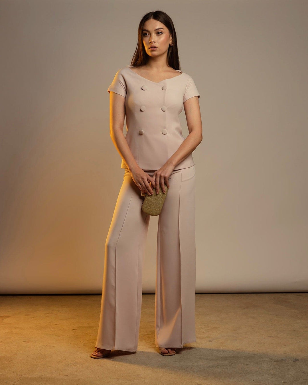 Woman wearing elegant beige pantsuit with wide-leg trousers and gold clutch in studio.