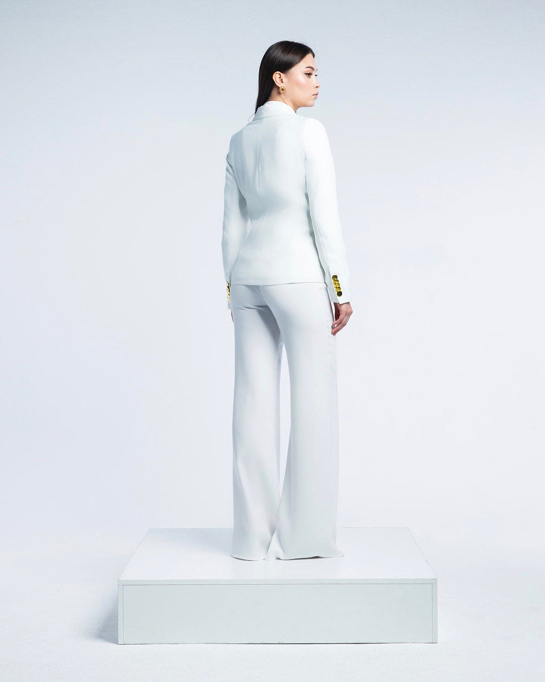 Woman modeling a white tailored suit with gold button details, standing on a white platform
