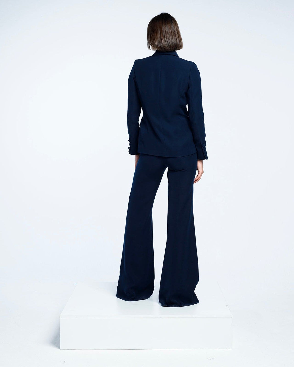 Woman in navy blue tailored suit with wide-leg pants, standing on white platform, back view