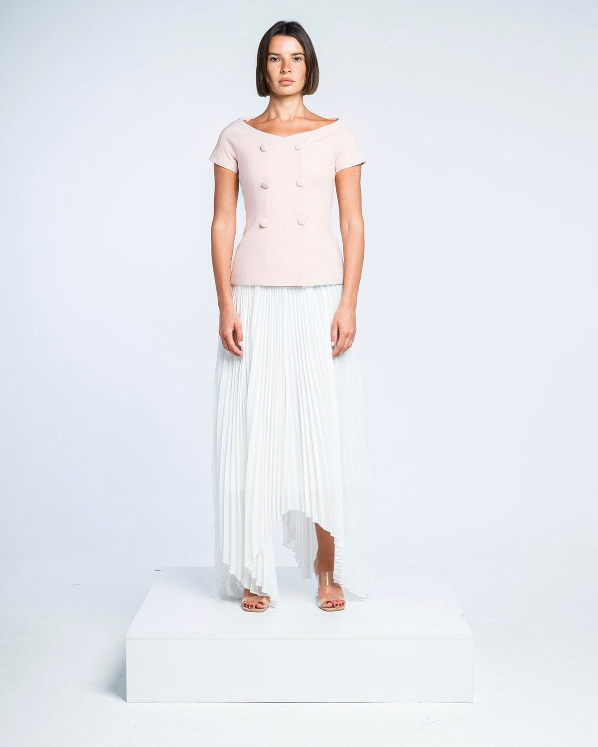 Woman modeling elegant blush pink top with button details and white pleated asymmetrical skirt