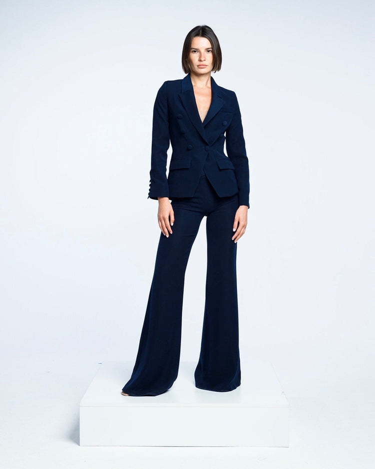 Woman in a tailored dark navy suit with flared pants, standing on a white platform against a plain background.
