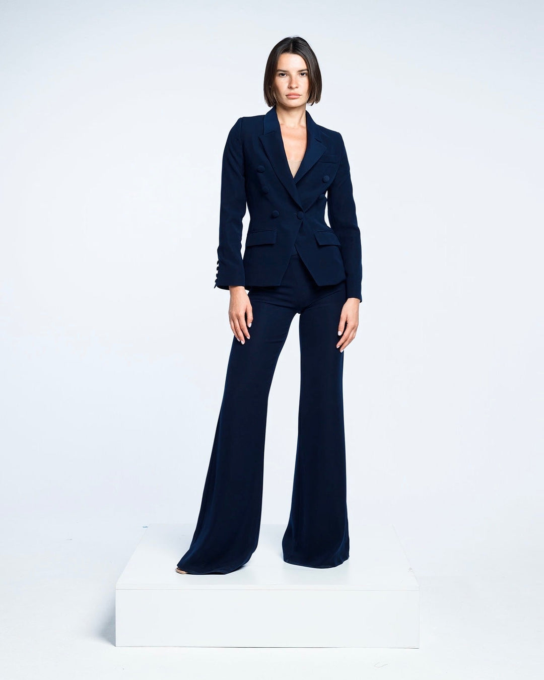 Woman in navy tailored suit with flared pants posing on white platform, minimalist background