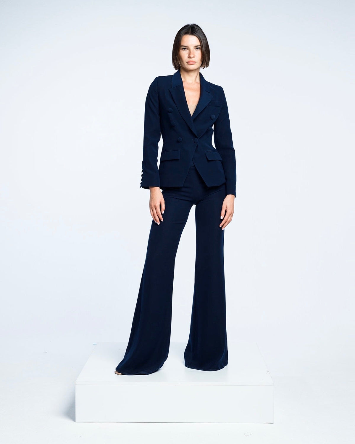 Woman in navy tailored suit with flared pants posing on white platform, minimalist background