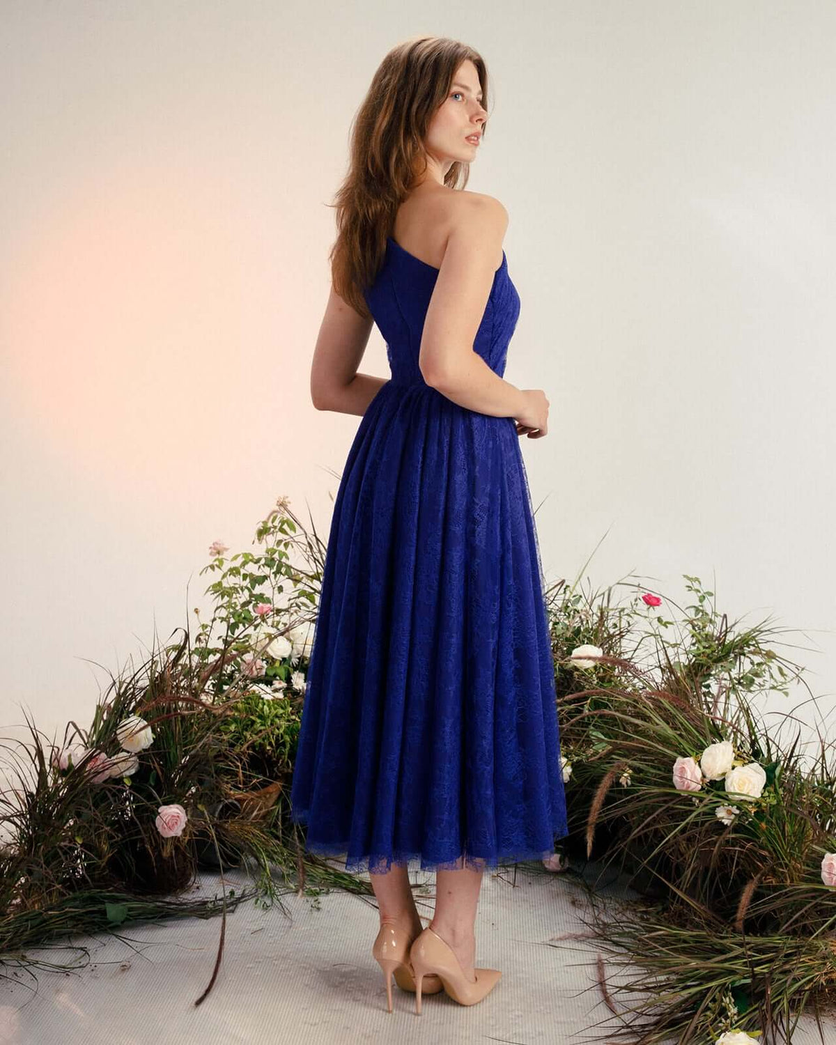Woman in elegant blue midi lace dress and nude heels standing among flowers