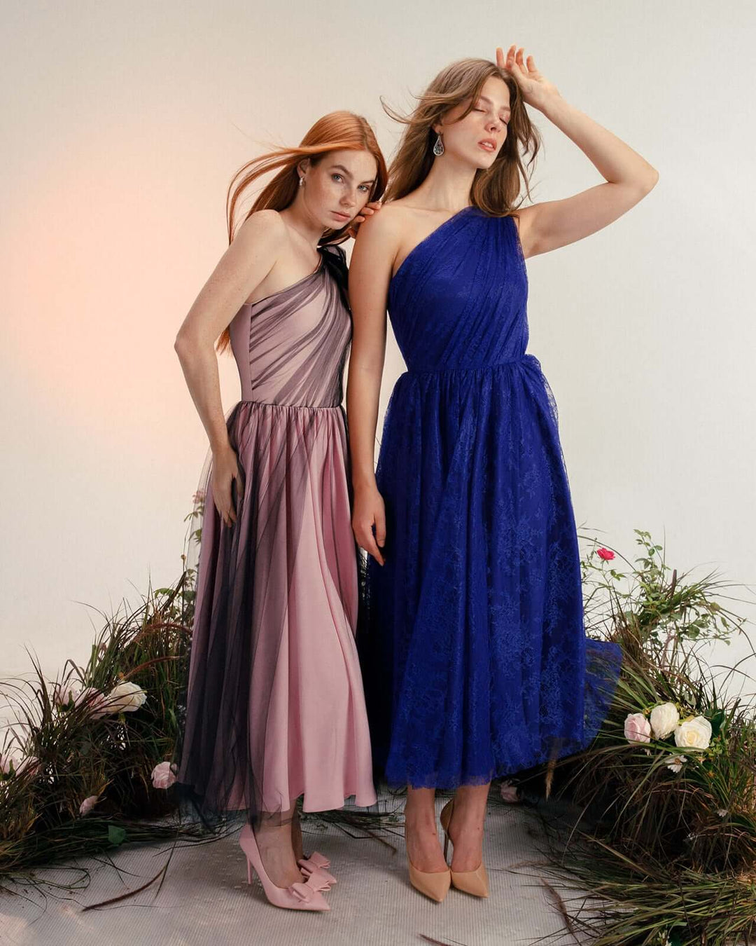 Two women wearing elegant midi evening dresses, one in pale pink tulle and one in royal blue lace, posing among flowers and foliage against a neutral backdrop.