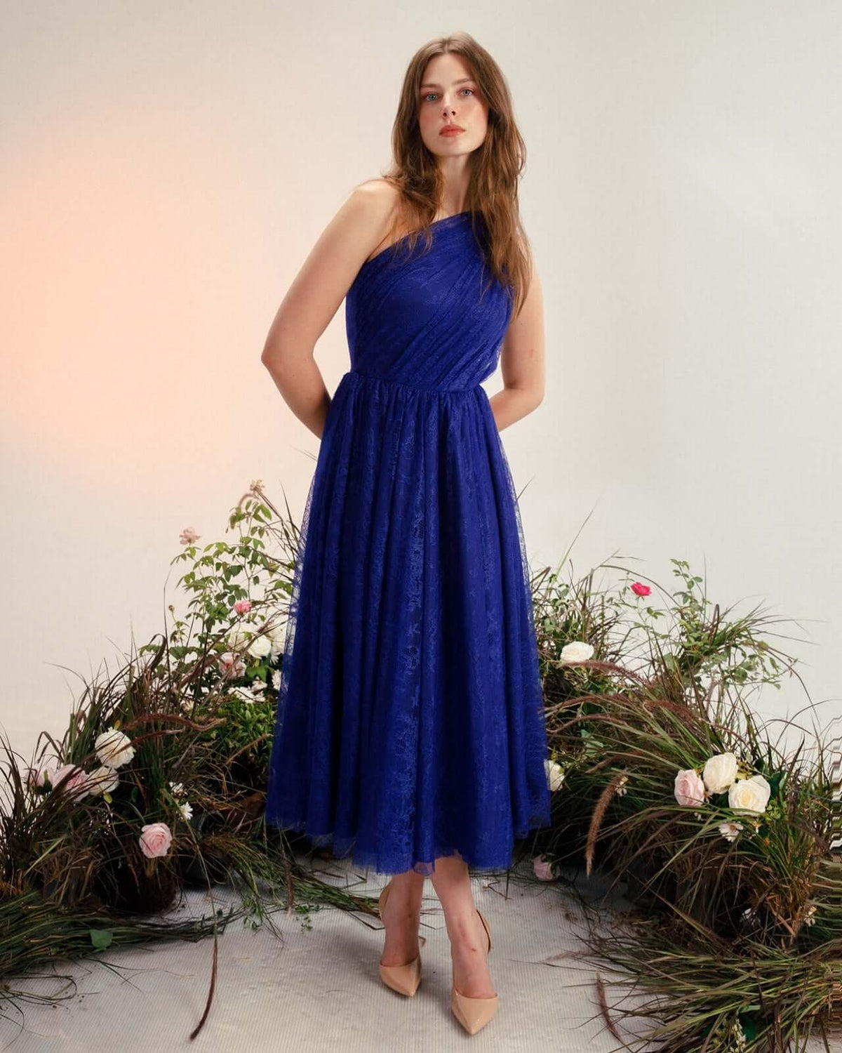 Woman in an elegant blue one-shoulder lace dress posing among flowers