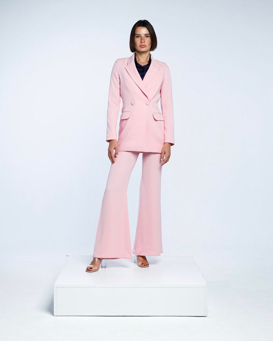 Woman in light pink tailored suit and flared pants posing on white platform, studio background