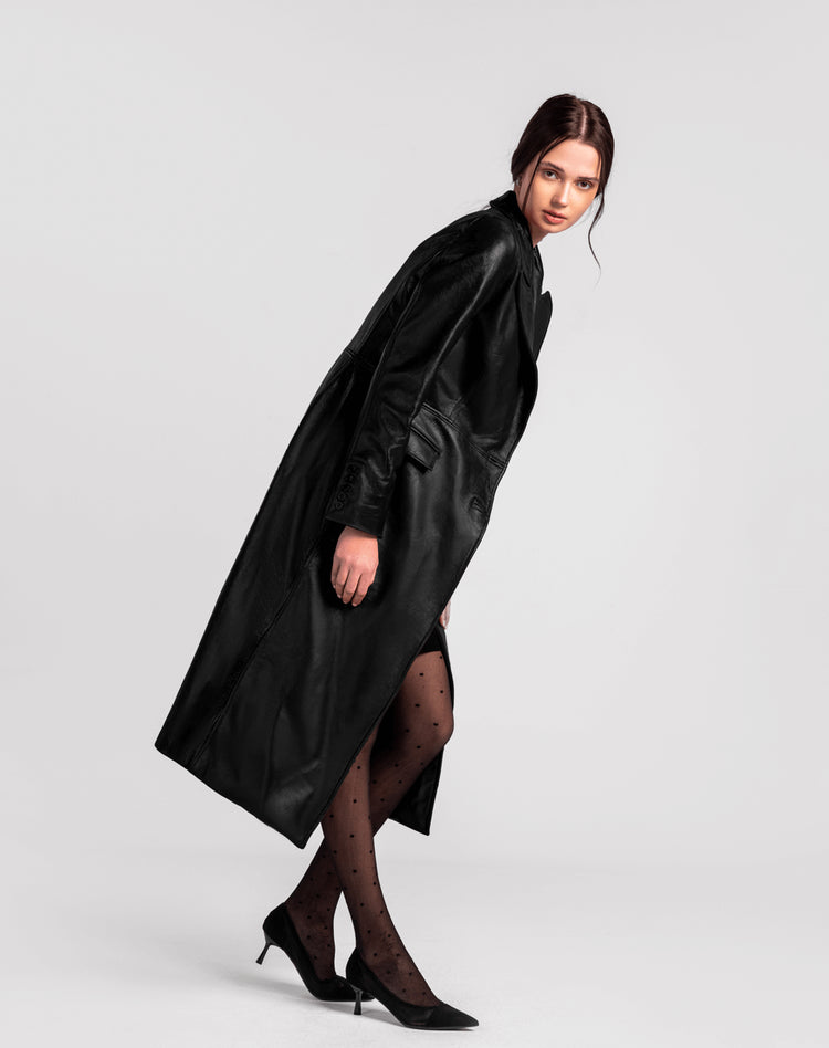 Woman in long black leather trench coat, sheer polka dot tights, and black heels, studio background.