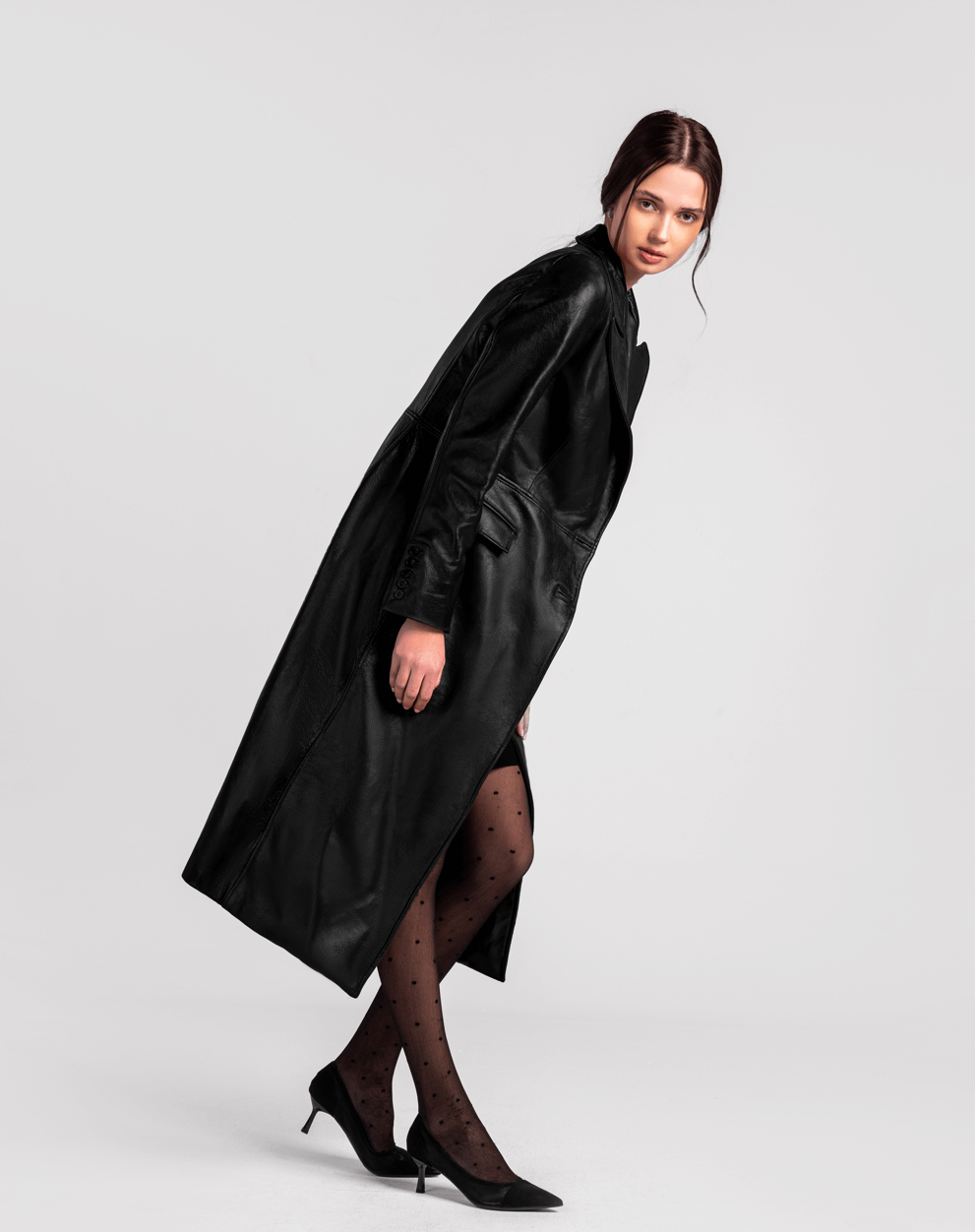 Woman in long black leather trench coat, sheer polka dot tights, and black heels, studio background.